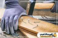 تركز على عملية تثبيت الألواح الخشبية يدوياً باستخدام المطرقة والمسامير. يظهر العامل وهو يرتدي قفازات الحماية